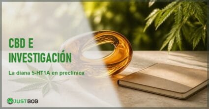 Bodegón sereno con una escultura abstracta de cristal ámbar translúcido, una hoja de cáñamo y un cuaderno cerrado sobre piedra crema, con follaje verde