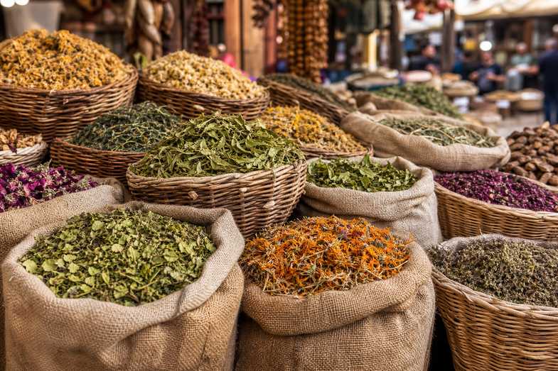 mercado-sacos-cestas-plantas-secas Puesto de mercado tradicional con sacos de arpillera abiertos y cestas de mimbre llenas de plantas secas de tonos verdes, ocres y anaranjados