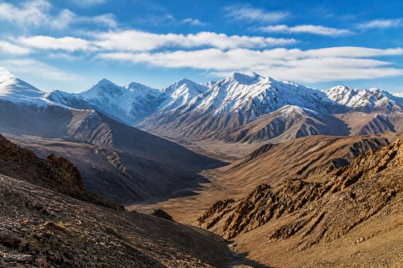 cordillera-alta-montana-valle-arido Paisaje de alta montaña con laderas áridas de tonos ocres, un valle profundo y cumbres nevadas al fondo bajo un cielo azul con nubes